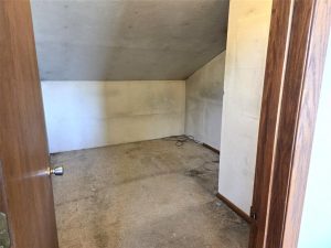 Small, empty room with slanted ceiling, stained carpet, and discolored white walls. A coiled black cable lies on the floor near the back wall. The room is seen through an open wooden door.