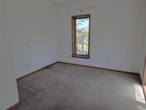 Empty room with light-colored walls, brown carpet, wood trim, and a single window letting in sunlight. Some electrical cords are on the floor near the wall. The space appears clean and undecorated.