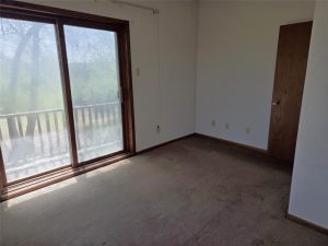 A small, empty room with brown carpet, white walls, a wooden door, and a large sliding glass door leading to a balcony with trees visible outside. Sunlight is streaming in through the glass door.