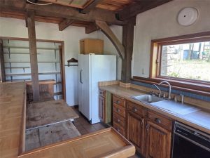 Rustic kitchen with wooden cabinets and countertops, a white refrigerator, open shelving, a double sink under a window, and exposed beams. There is a built-in wooden table with bench seating.