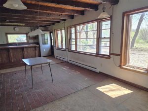 Sunlit room with large windows, exposed wooden beams, and a brick floor. A small table sits in the center, and there is a bar area in the background. The room appears empty and rustic, with views of trees outside.