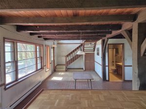 A rustic, sunlit interior features exposed wooden beams, large windows, a wooden staircase, and a small table on a tiled floor. A bathroom with a sink and mirror is visible through an open door on the right.