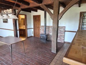 A rustic room with brick tile flooring, exposed wooden beams, a wood-burning stove on a brick platform, a folding table, and a wooden bar counter; a bathroom with a vanity is visible through an open door.