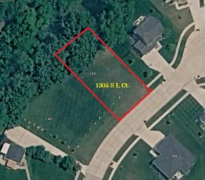 Aerial view of a residential neighborhood showing an empty rectangular lot outlined in red, labeled "1305 S L Ct." The lot is surrounded by houses, driveways, and a wooded area.