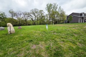 A grassy vacant lot with a "For Sale" sign in the center, bordered by trees in the background. Large rocks are on the left, and a two-story house is partially visible on the right. The sky is cloudy.
