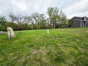 A grassy vacant lot with a "For Sale" sign in the center, bordered by trees in the background. Large rocks are on the left, and a two-story house is partially visible on the right. The sky is cloudy.