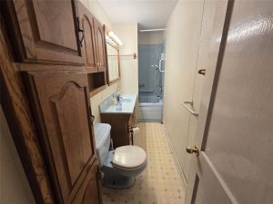 Narrow bathroom on with wood cabinets, a light blue sink and toilet, mirror above the sink, and a shower-tub combo with sliding glass doors. The floor features light patterned linoleum.