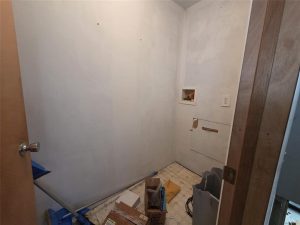 A small, unfinished laundry room with bare white walls, exposed pipes, a tile floor, and scattered boxes and supplies on the ground. A small wall outlet and laundry hookups are also visible.