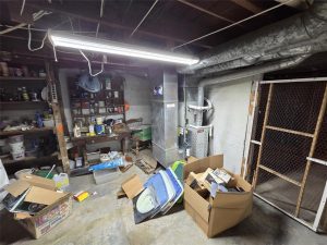 A cluttered basement with exposed pipes, shelves of tools and paint cans, a furnace, fluorescent lighting, and scattered boxes filled with various items on the concrete floor.