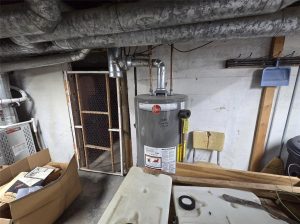 A gray Rheem water heater stands in the basement with exposed pipes overhead, surrounded by cardboard boxes, a broom, shovel, and a chair against the concrete wall.