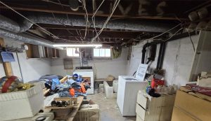 A cluttered basement laundry room with exposed pipes and wires, two washing machines, scattered clothes, shelves with jars, and various household items. The unfinished walls add to the messy appearance.