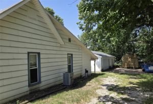 A light-colored house features a side window, central air unit, and gravel driveway surrounded by trees. In the background are a detached white garage, wooden shed, and blue dumpster under shade.