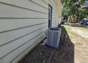 A gray outdoor air conditioning unit sits on a black platform next to the beige siding of a house with a grassy yard, shed, and blue dumpster visible in the background.