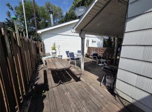 A wooden deck features a picnic table, potted plants, patio chairs, a small grill, and privacy fence. The deck is partly shaded by a roof attached to the white house.