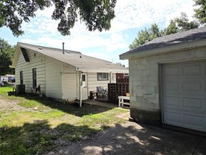 A charming small white house with a covered porch, separate garage, fenced patio, grassy yard, and shady trees under a partly cloudy sky.