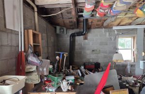 A cluttered garage with exposed insulation, hats hanging from the ceiling, assorted tools, boxes, and various items scattered on shelves and tables. A small window lets in some natural light.