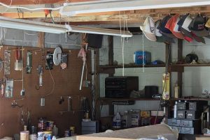 A cluttered garage workspace with tools hanging on a pegboard, shelves lined with cans, hats on a beam, and various items including boxes, a stereo, and drawers scattered around.