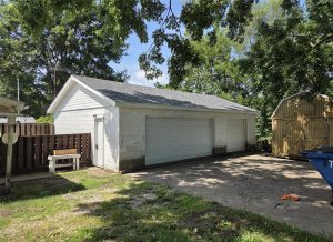 This white detached garage with a double door sits next to a small wooden shed. Surrounded by trees and a fenced yard, the property features a driveway out front and sunlight filtering through the branches—ideal real estate.