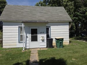 Charming small white house with gray roof. Labeled "403" above the door, it features a walkway flanked by bins, two windows, and is nestled among grass and trees.