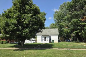A small white house with a gray roof sits behind a large leafy tree on a grassy lawn. A sidewalk runs in front, and several tall trees and a white van are visible nearby under the blue sky.