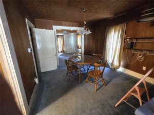 A small dining room with wood-paneled walls, four wooden chairs around a round table, a chandelier above, carpeted floor, old white door, long curtains, shelves on the wall, and an adjacent room visible.