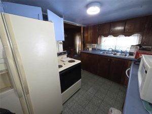 Small, dimly lit kitchen with dark brown cabinets, blue countertops, white appliances including a fridge, stove, and microwave, green tiled floor, and a window with sheer curtains above the sink.