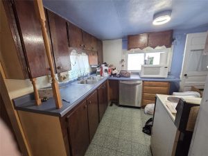 A small kitchen features dark wood cabinets, blue countertops, a double sink, dishwasher, and microwave by the window. The green-tiled floor and scattered items add a lived-in charm.