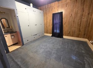 A small room features wood-paneled walls, blue carpet, a window with black curtains, built-in white storage cabinets, and an open doorway to a bathroom with a sink and mirror.