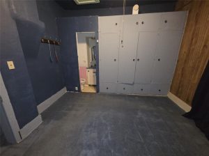 A small, empty room in Indianola with dark blue walls and carpet, a wall-mounted coat rack, built-in light blue cabinets, and an open door leading to a bathroom with visible toilet and sink
