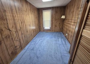 A small, empty room features wood-paneled walls, blue carpet, a single window with closed blinds, a wall-mounted lamp, and an exposed black cable hanging from the left wall—ideal for real estate photos.